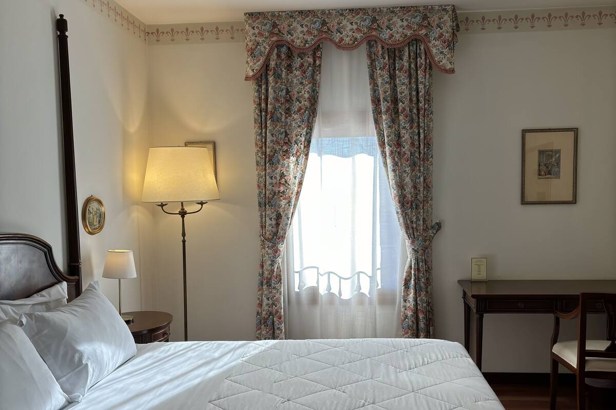 A neatly made bed with a quilted cover, a bedside lamp, a small round table with a lamp, a desk with a chair, and a window with floral curtains.