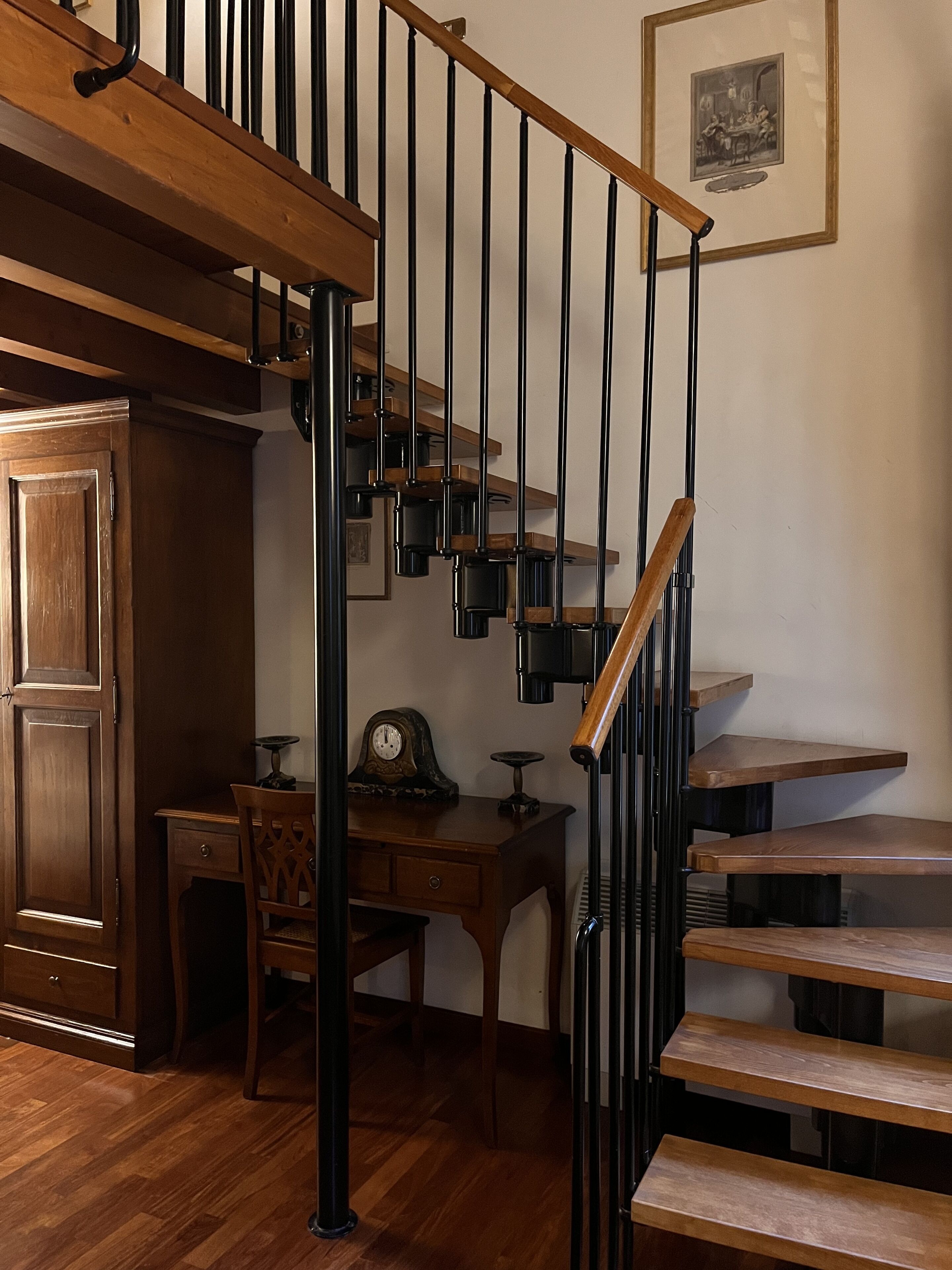 Una escalera de madera con barandillas de metal negro, una mesa de comedor con sillas y un reloj fijado a la pared.