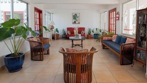 Amplia sala de estar con un sofá rojo, sillas de madera, una mesa de centro y diversas plantas en macetas.