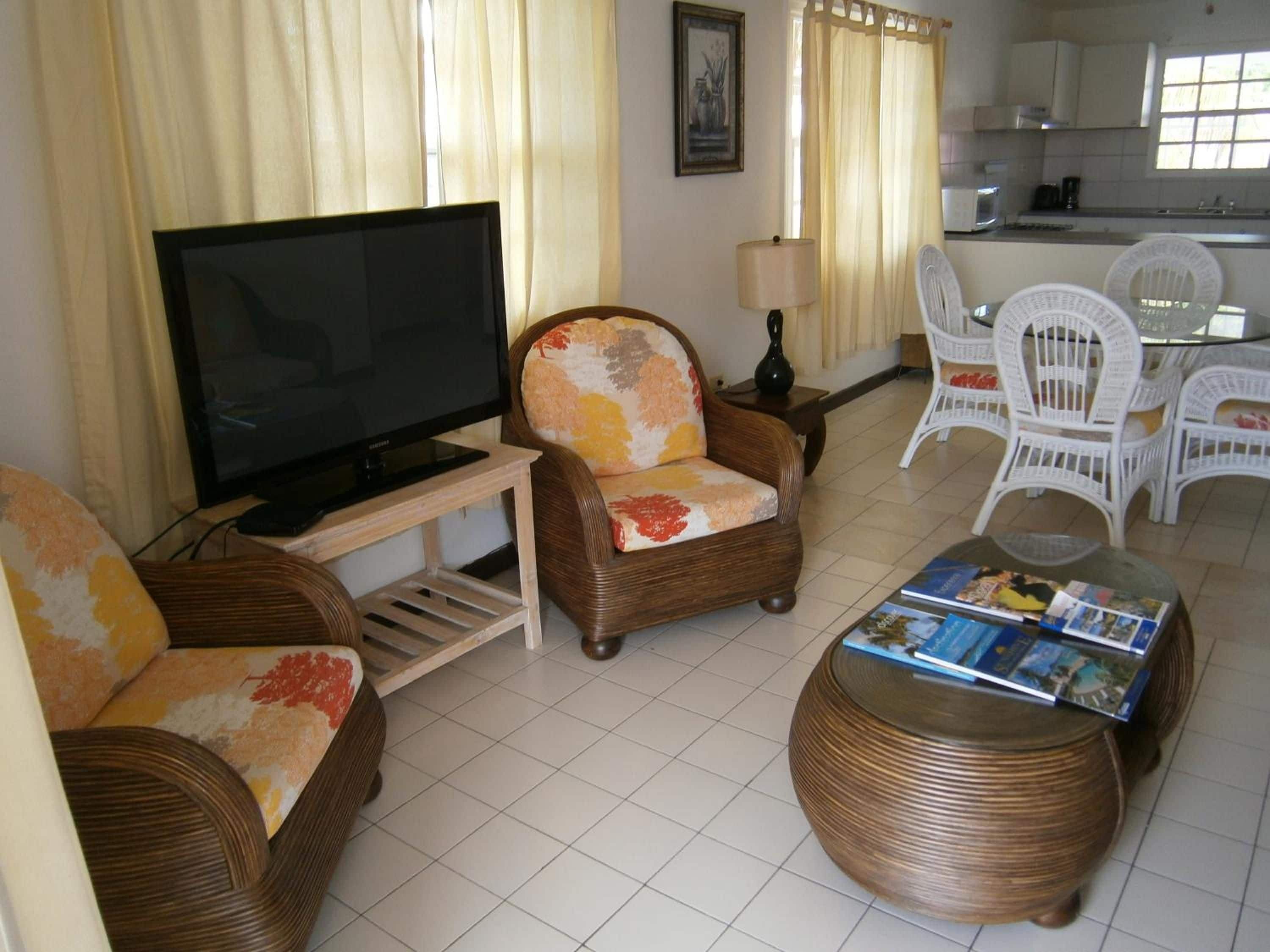 Una sala de estar con televisor, muebles de mimbre, una mesa de centro con revistas y una cocina al fondo.