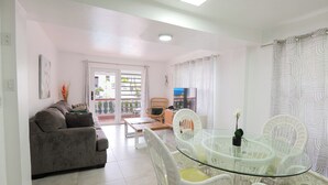 Una sala de estar con un sofá, una mesa de vidrio con sillas, un televisor y un balcón con plantas.
