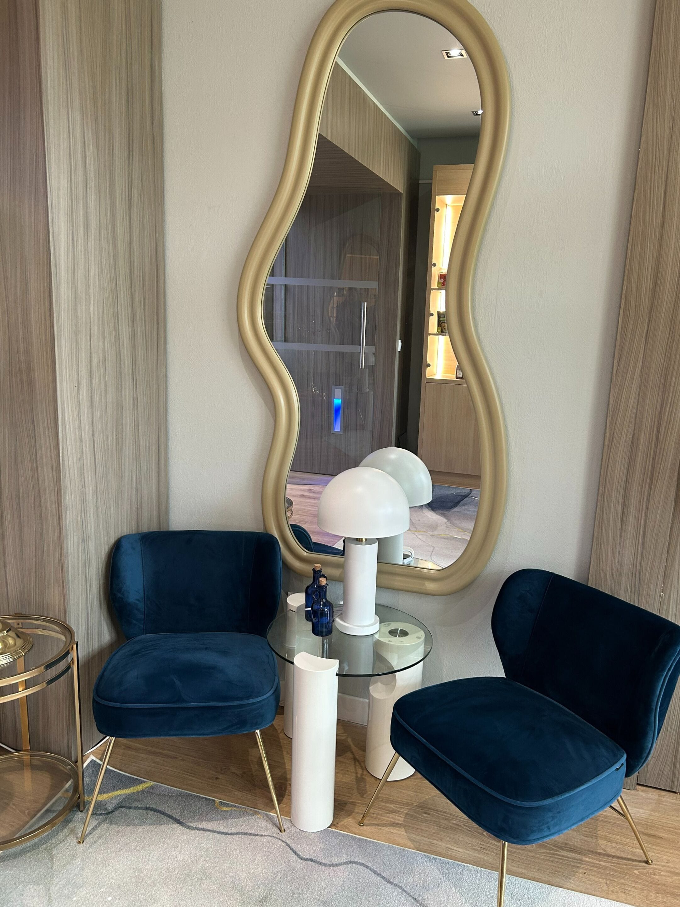 A modern living room with two blue armchairs, a glass coffee table, a white lamp, and a large mirror with a gold frame.