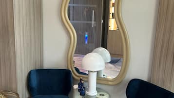 A modern living room with two blue armchairs, a glass coffee table, a white lamp, and a large mirror with a gold frame.