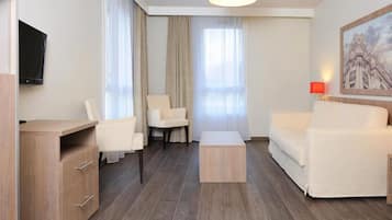 A modern hotel room with a wooden desk, two white armchairs, a white sofa, a red pendant light, and a painting of a building on the wall.