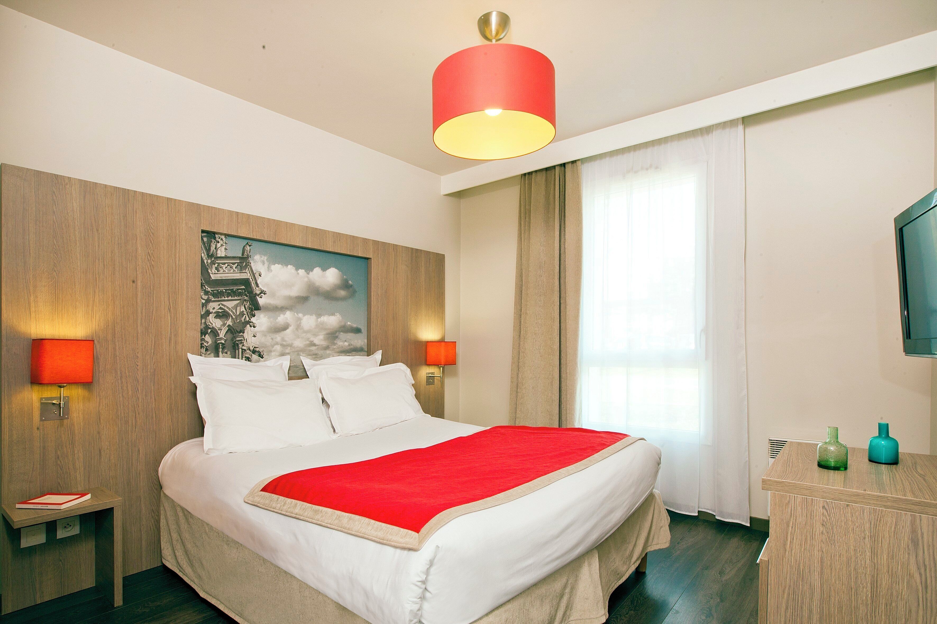 Habitación de hotel con cama, dos lámparas de noche, lámpara de techo roja, cabecero de madera, un cuadro, ventana con cortinas, cómoda de madera y televisión.