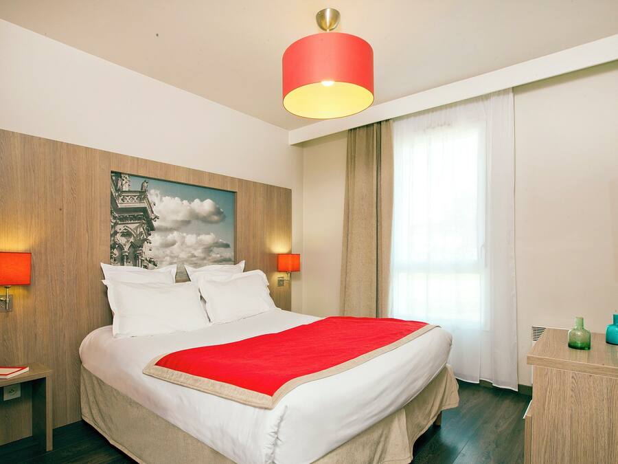 Habitación de hotel con cama, dos lámparas de noche, lámpara de techo roja, cabecero de madera, un cuadro, ventana con cortinas, cómoda de madera y televisión.