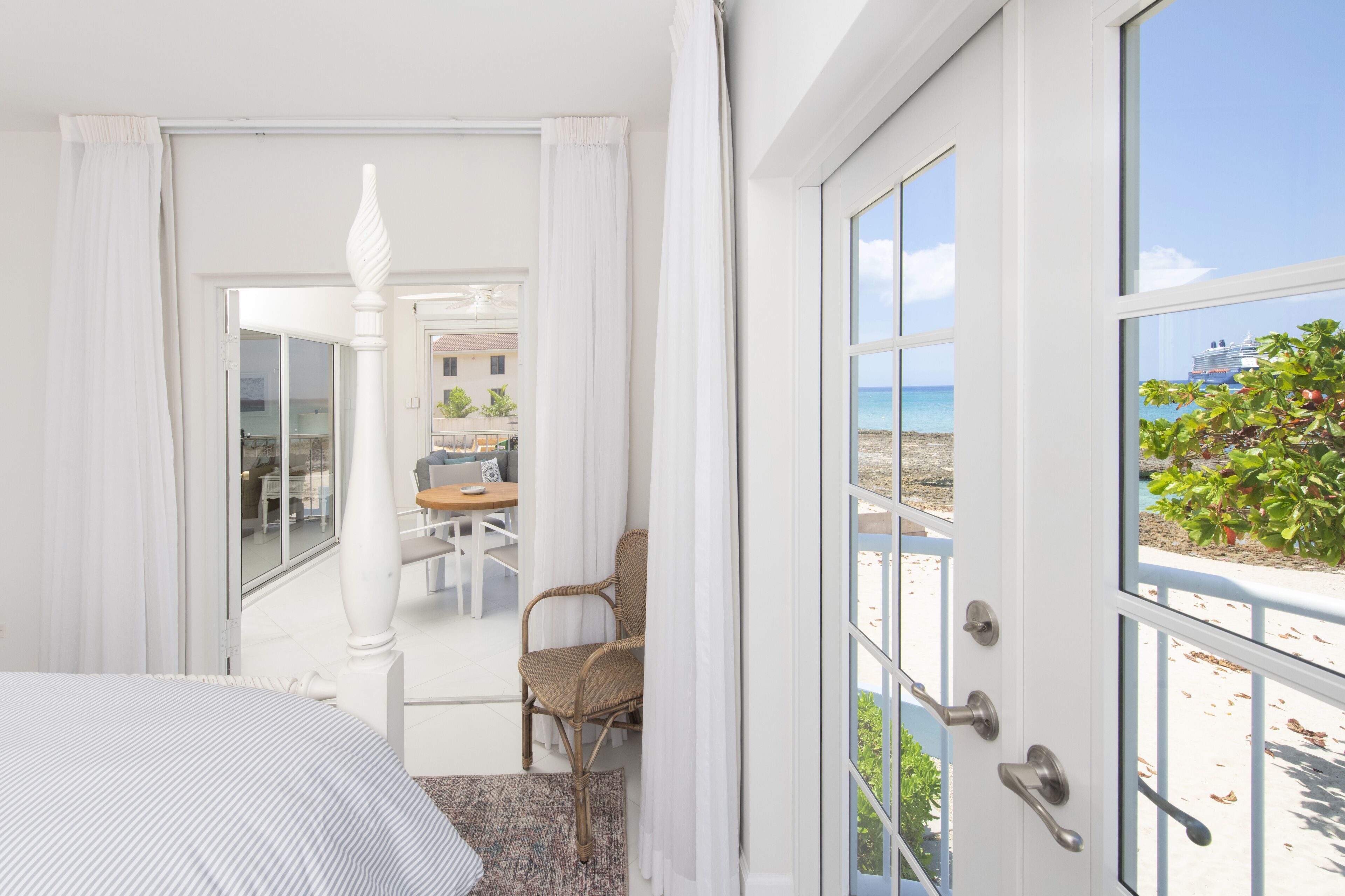 Una habitación de hotel con una cama, una silla de mimbre, una zona de comedor con mesa y sillas, y vistas a la playa y al océano a través de amplios ventanales.