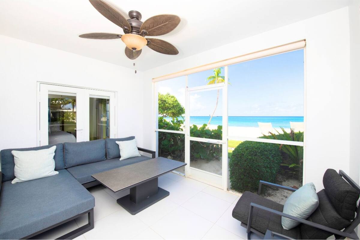 A modern living room with a sofa, a coffee table, and a chair. There is a ceiling fan and a large window with a view of the beach.