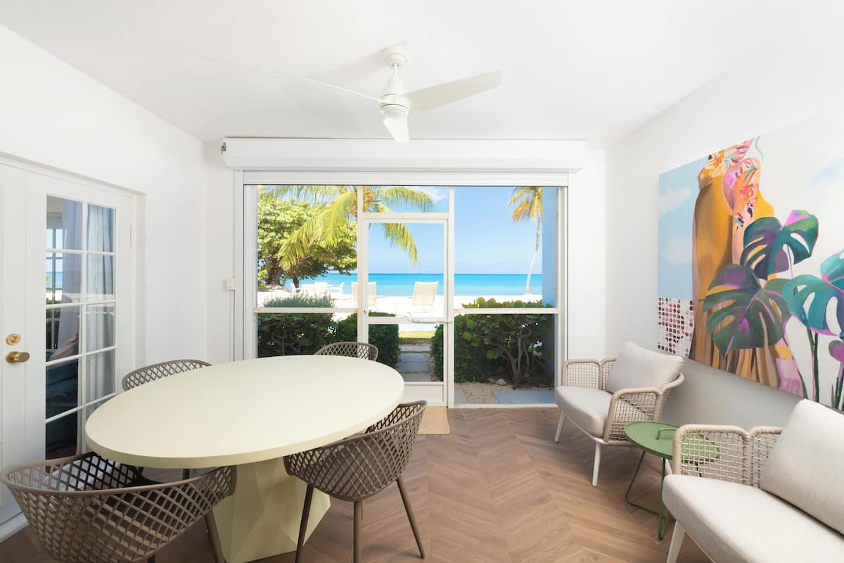 A room with a dining table and chairs, a sofa, a ceiling fan, and a view of the ocean through large glass doors.