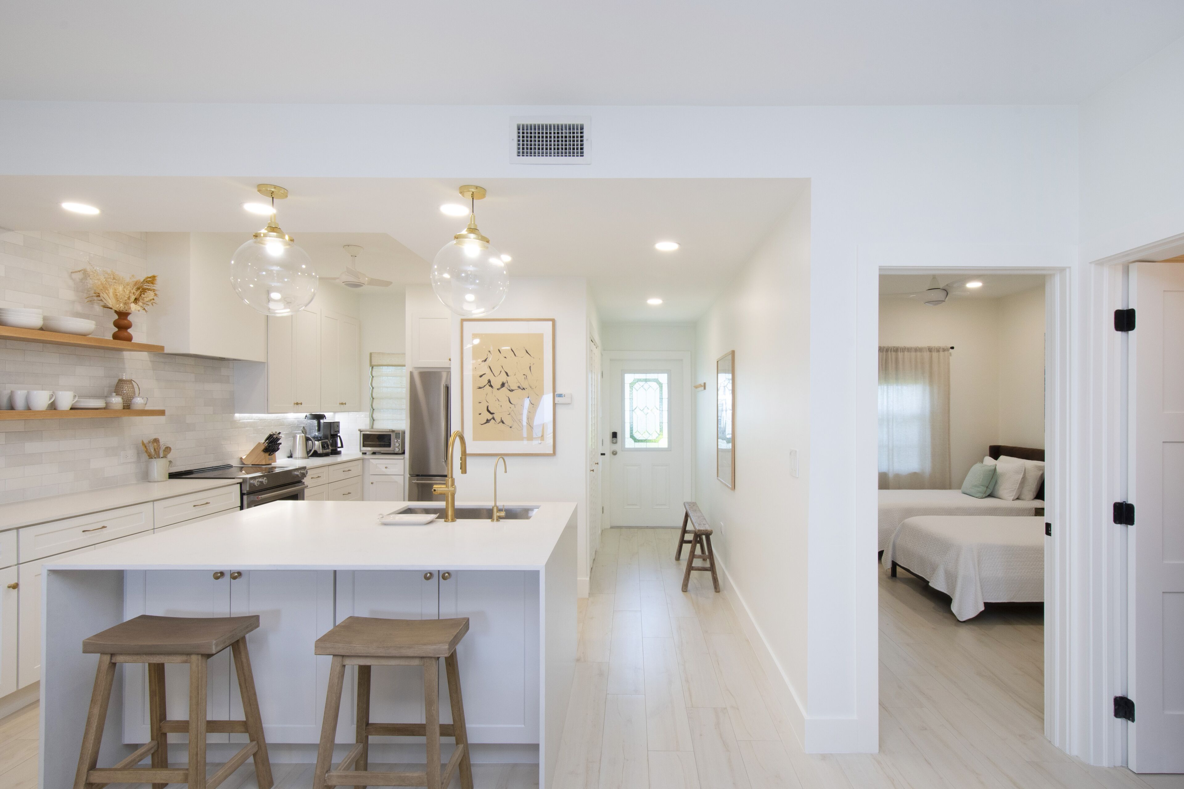 A modern kitchen with white cabinetry, a large island with stools, pendant lighting, and a view into a bedroom with two beds.