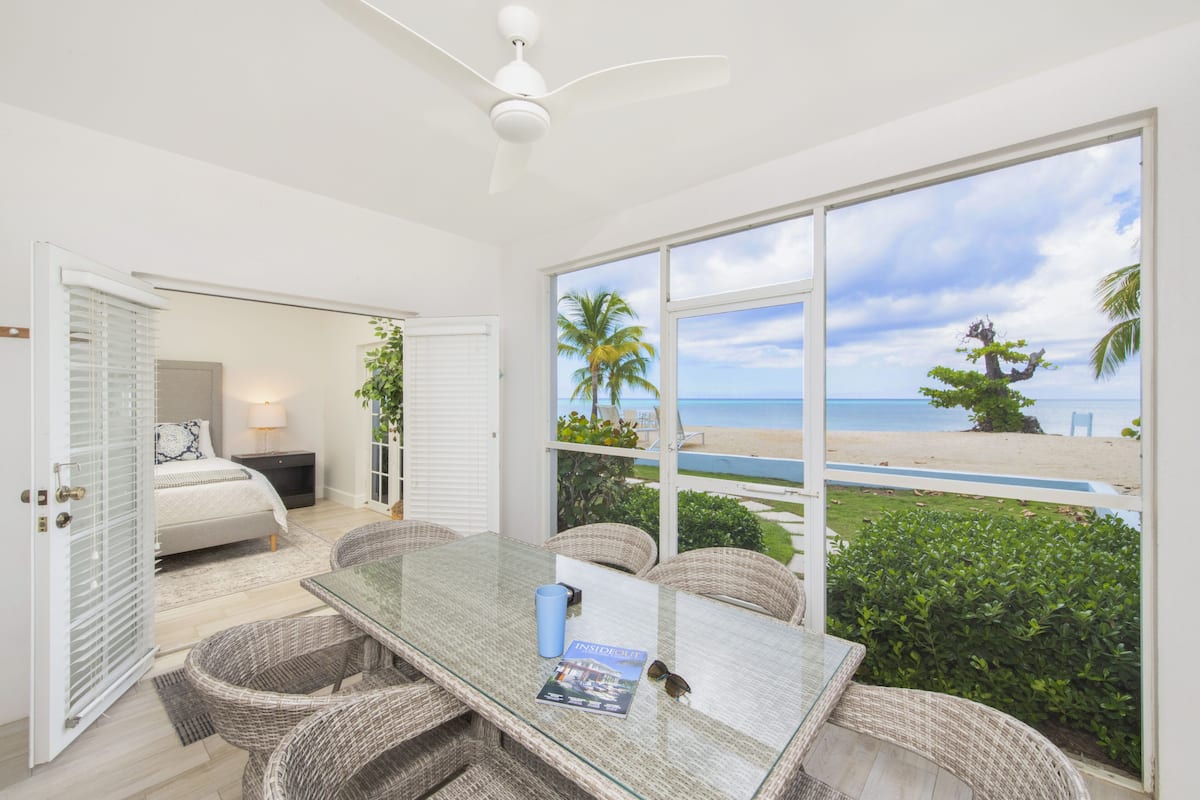 A room with a glass table, wicker chairs, a bed, and a view of the beach.