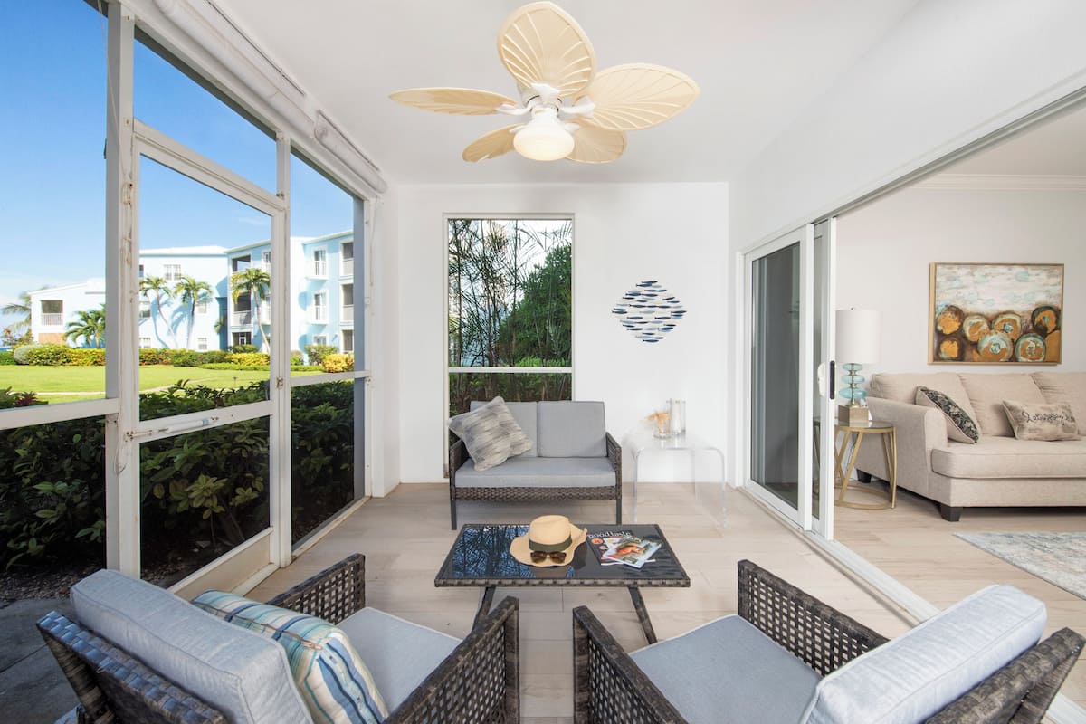 A spacious balcony with a ceiling fan, a sofa, a coffee table, and a view of a building and greenery.