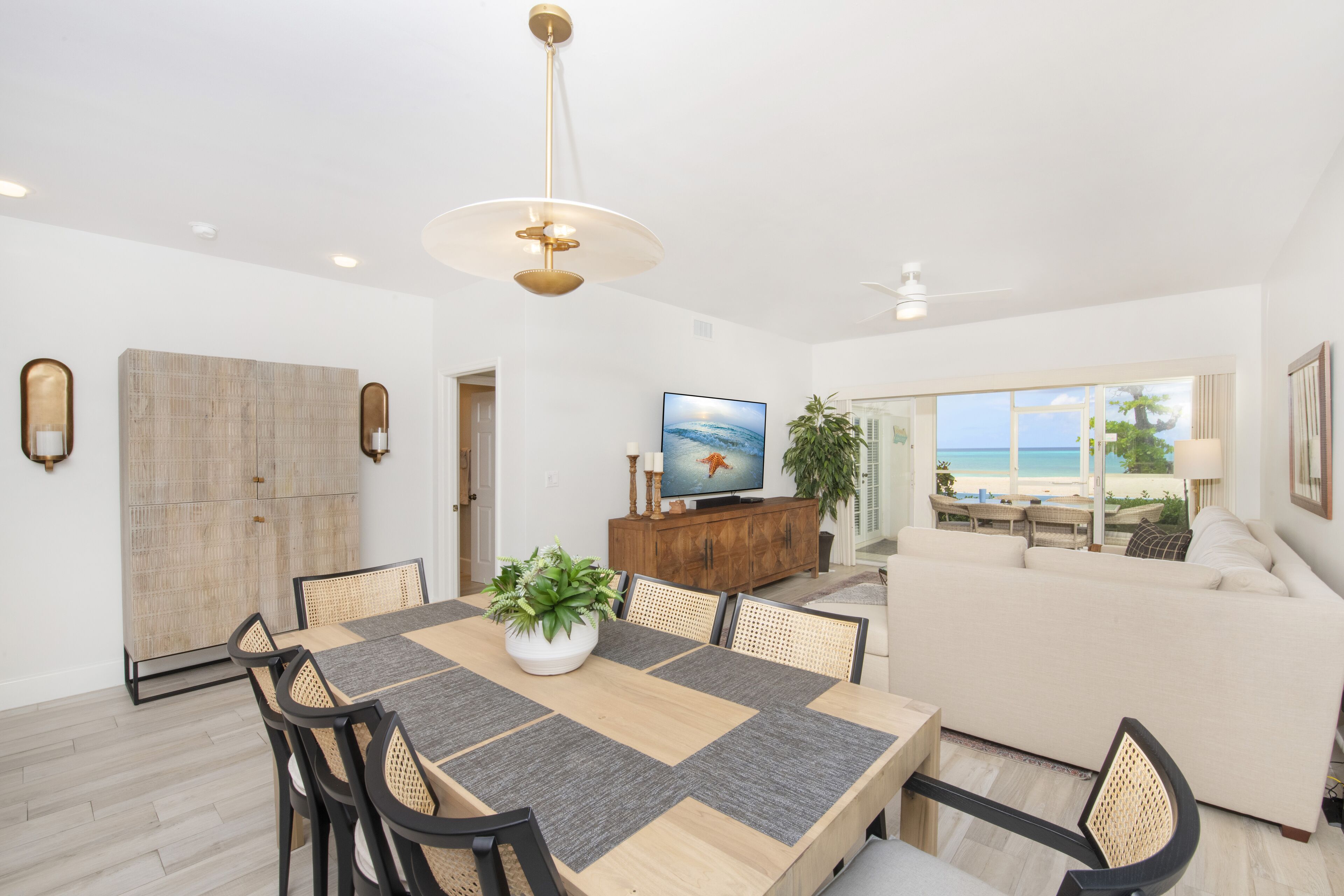 Amplia sala de estar con un ventanal que ofrece vistas a la playa, comedor con mesa y sillas de madera, y un rincón de descanso acogedor con sofá y cojines.