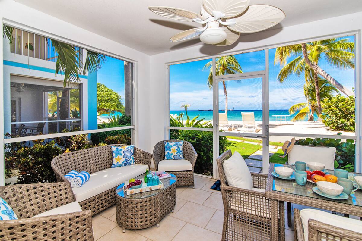 A room with wicker furniture, a glass coffee table, and a view of a beach.