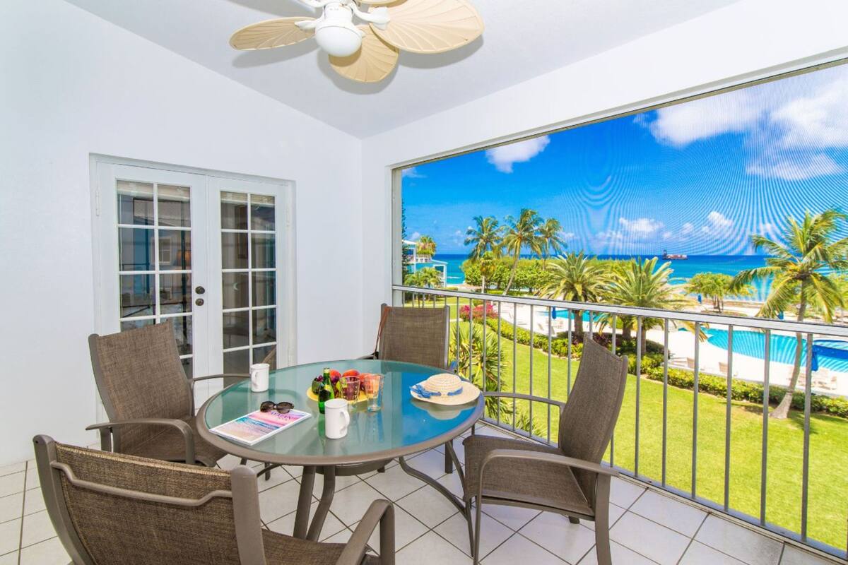A balcony with a glass table, chairs, and a view of a beach and pool.
