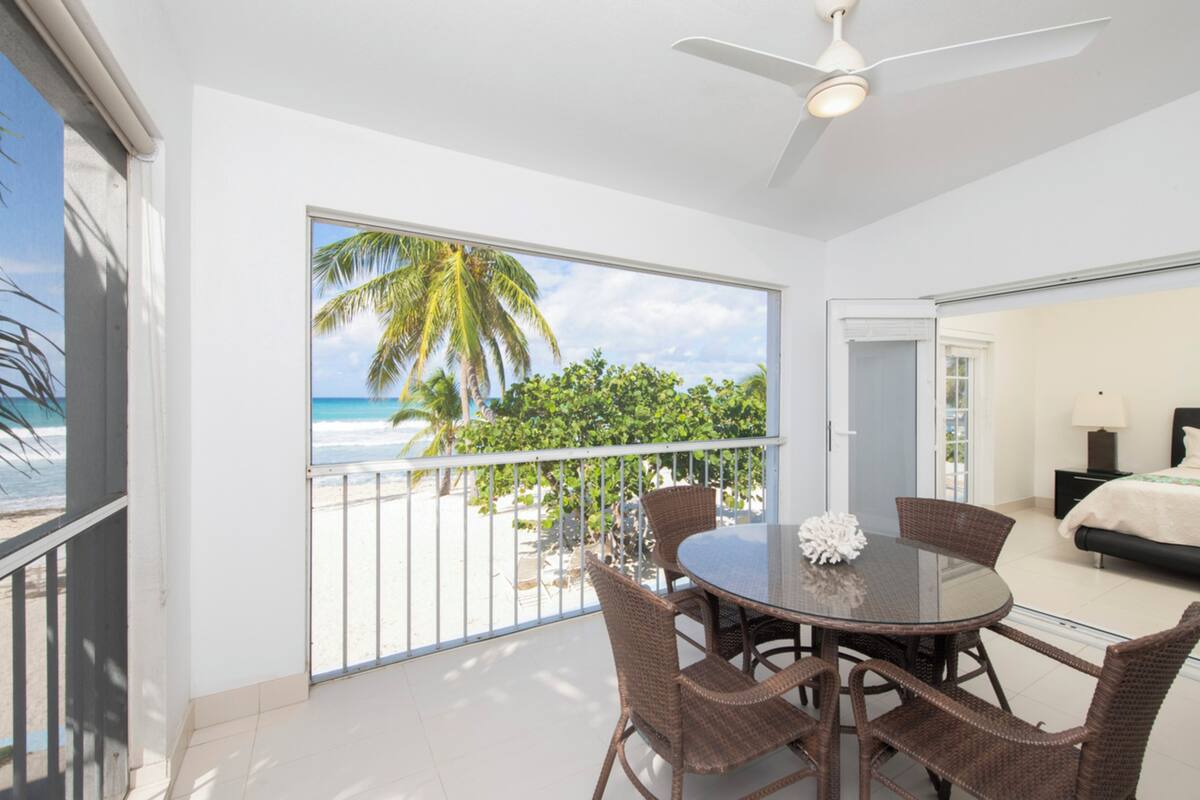 A balcony with a view of the beach and palm trees, a dining table with wicker chairs, and a ceiling fan.