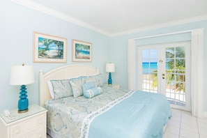 A bedroom with a bed, two nightstands, a door leading to a balcony, and two framed paintings on the wall. - The Grandview Condos on Seven Mile Beach (Seven Mile Beach)