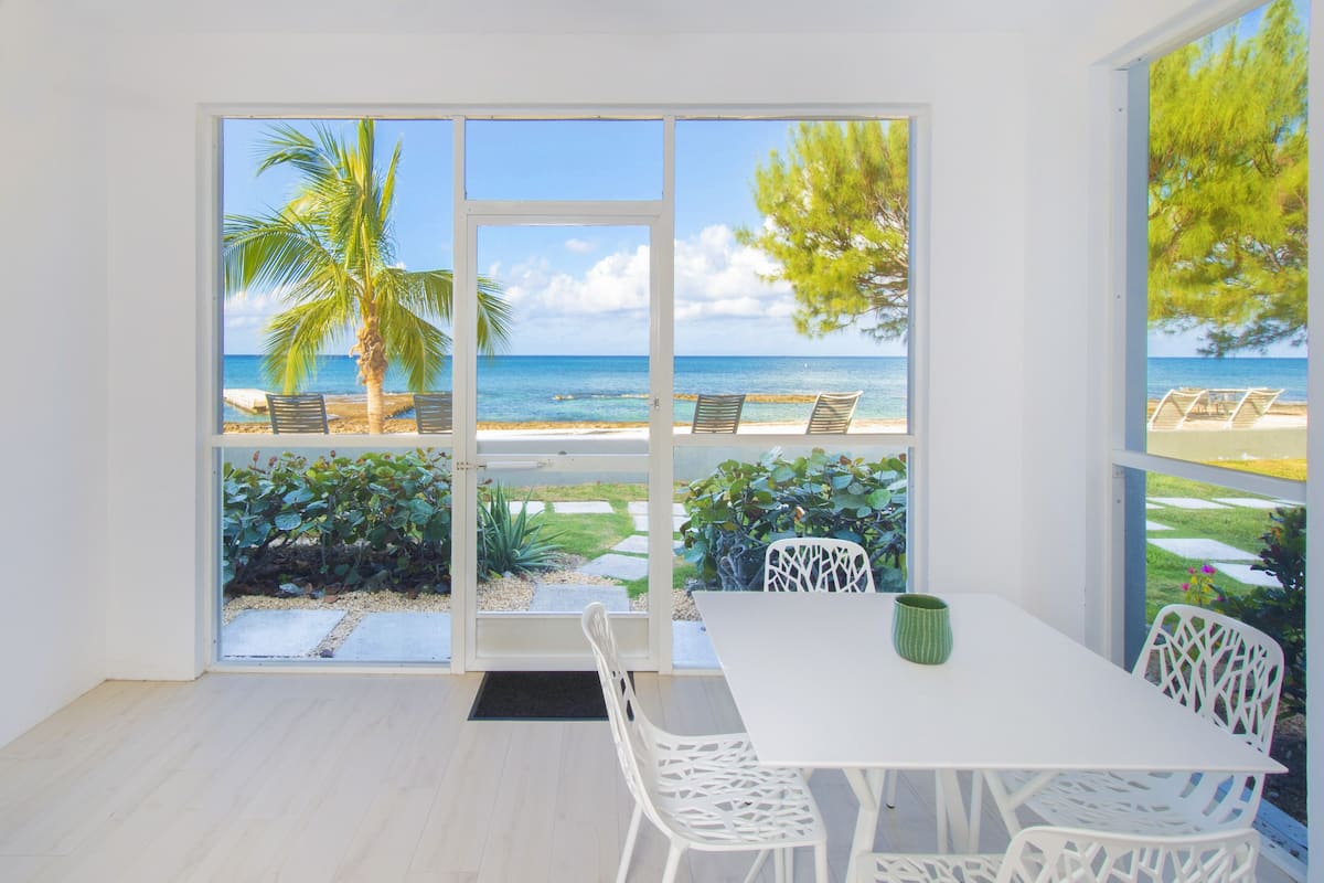 A room with a white table and chairs, a green vase, and a view of a beach with palm trees and a clear sky.