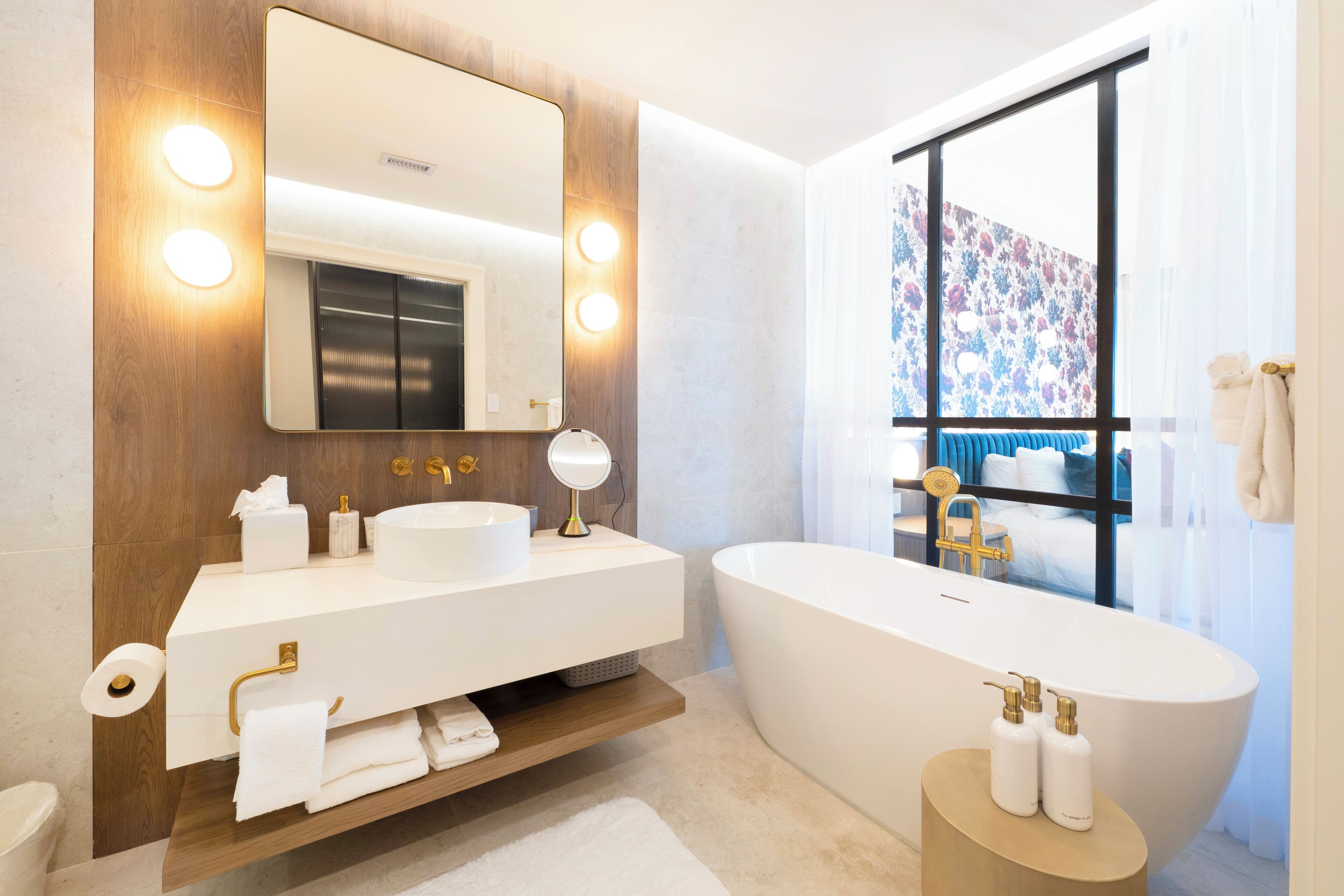Un baño moderno con un espejo grande, dos lavamanos, una bañera independiente y un ventanal con vistas a una cama y cortinas.