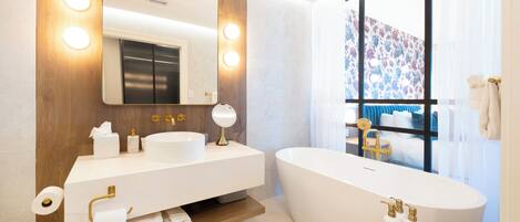 Un baño moderno con un espejo grande, dos lavamanos, una bañera independiente y un ventanal con vistas a una cama y cortinas.