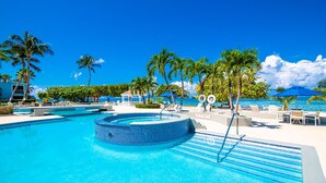 A clear blue swimming pool with a circular hot tub, surrounded by lounge chairs and palm trees, with a view of the ocean. - The Grandview Condos on Seven Mile Beach (Seven Mile Beach)