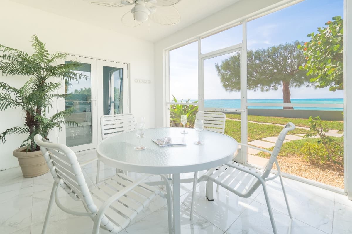 A bright room with a round table and chairs, large windows with a view of the sea, and a potted plant.