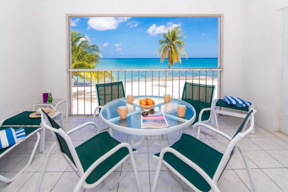 A round table with chairs, a magazine, and a bowl of fruit on a balcony with a view of the beach and palm trees.