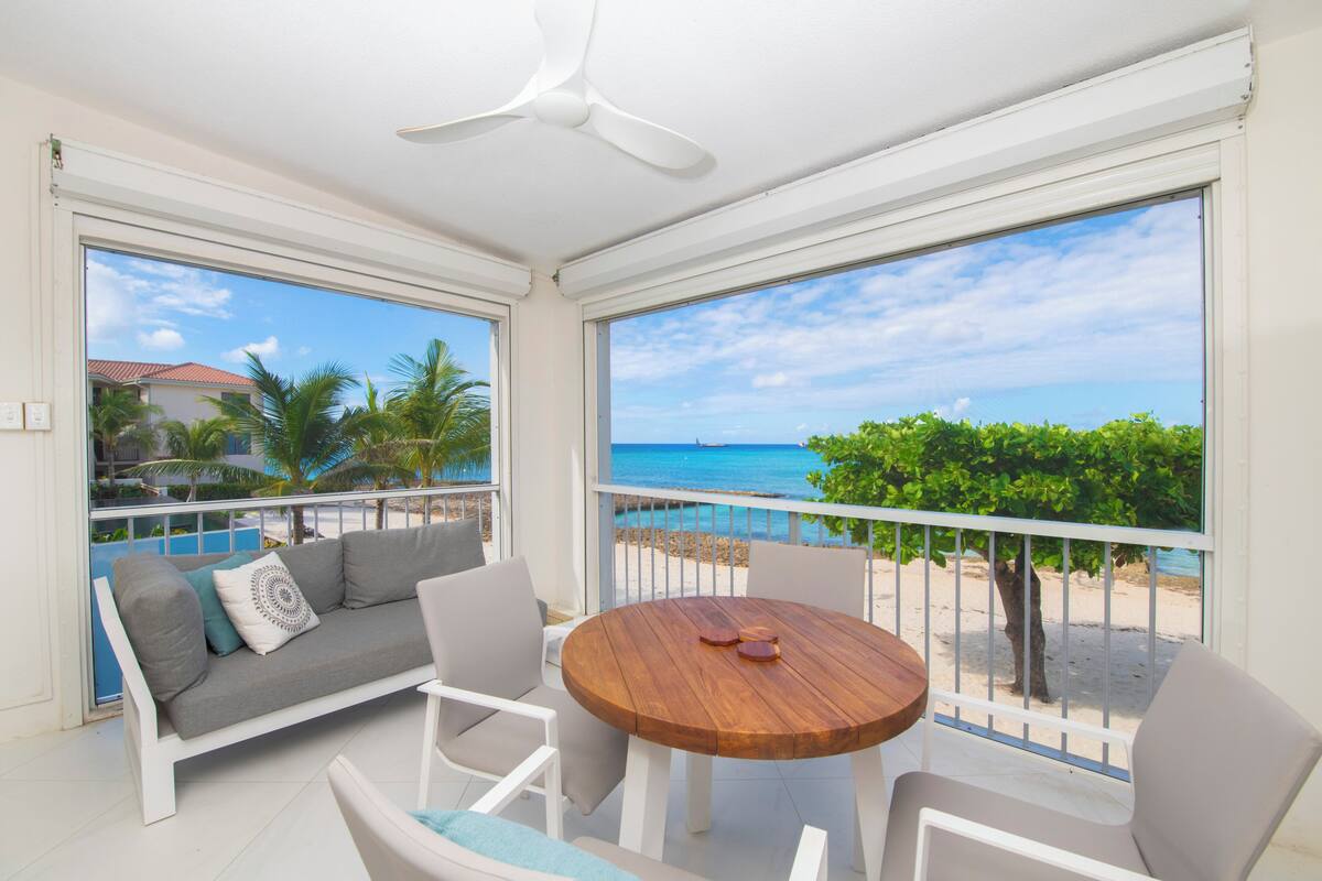 A balcony with a white sofa, a wooden table, and chairs, overlooking a beach and ocean.