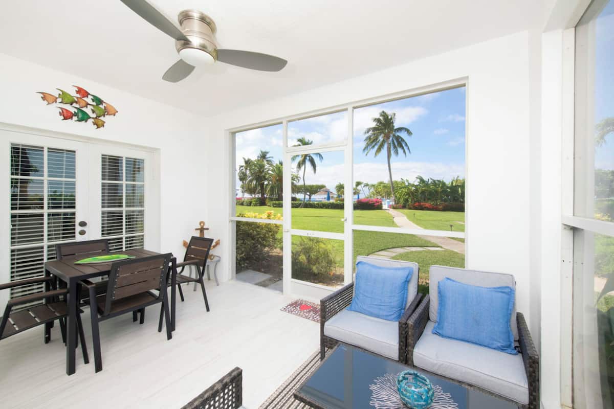 A room with a dining table and chairs, a sofa with cushions, a ceiling fan, and large windows offering a view of a garden with palm trees and a pathway.