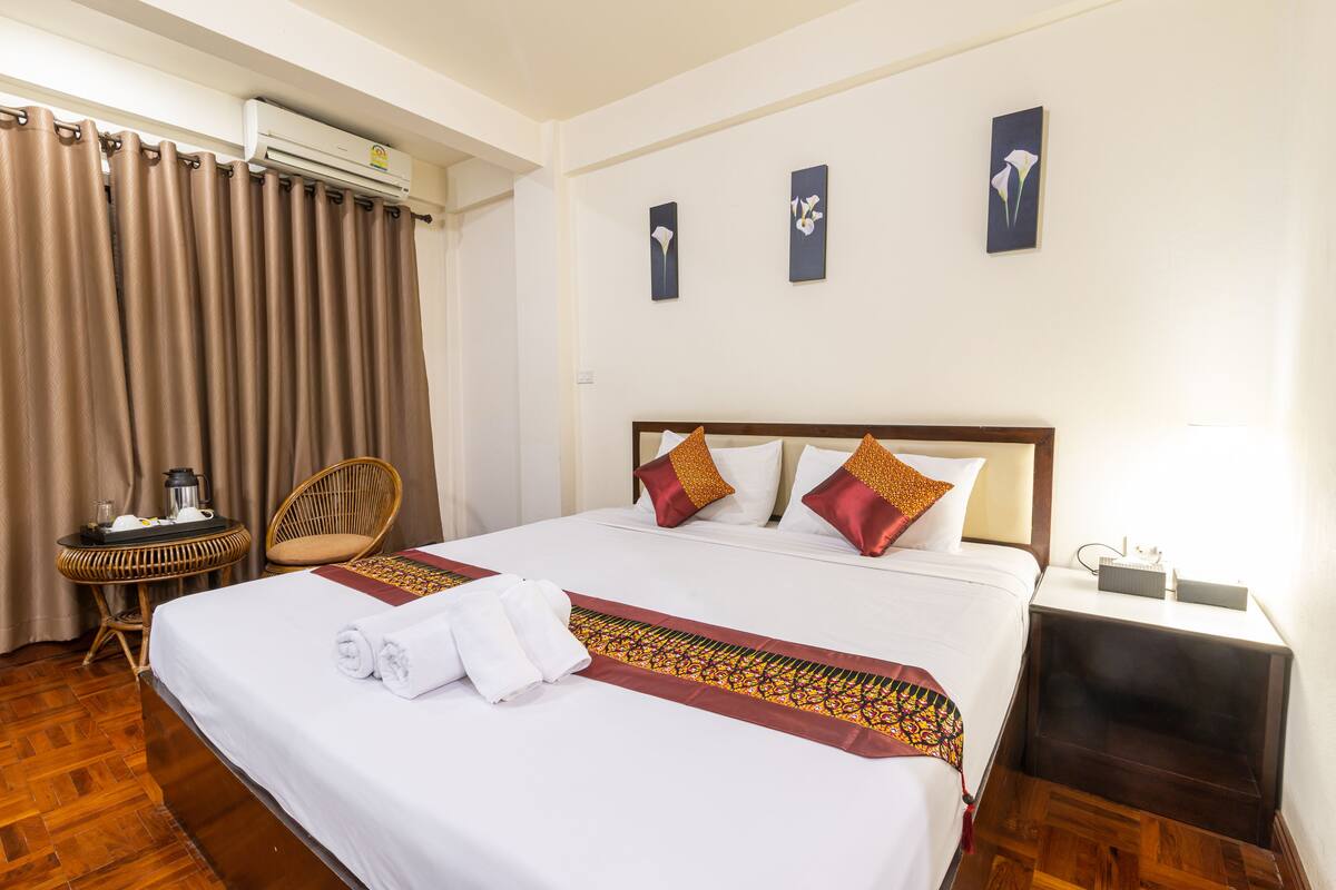 A hotel room with a large bed, a bedside table, a chair, and a wall-mounted air conditioner.