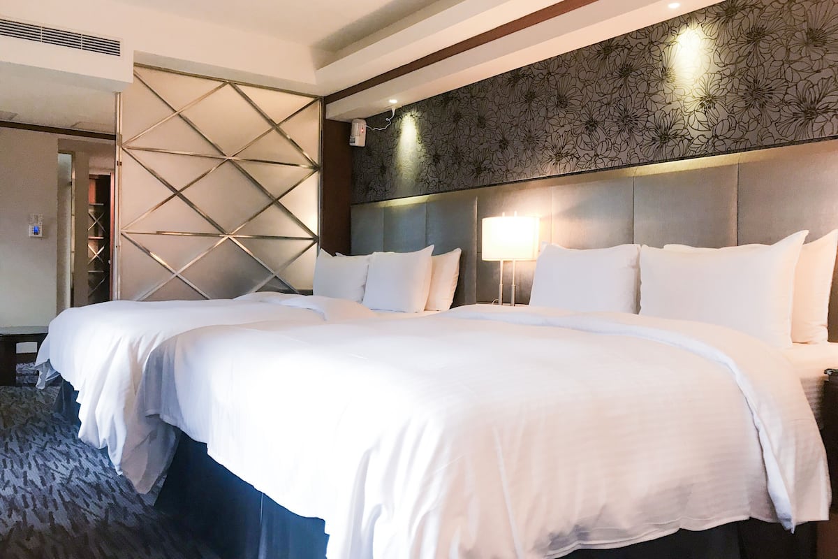 A hotel room with a large bed, a headboard with a floral pattern, a wall-mounted air conditioner, and a patterned wall.