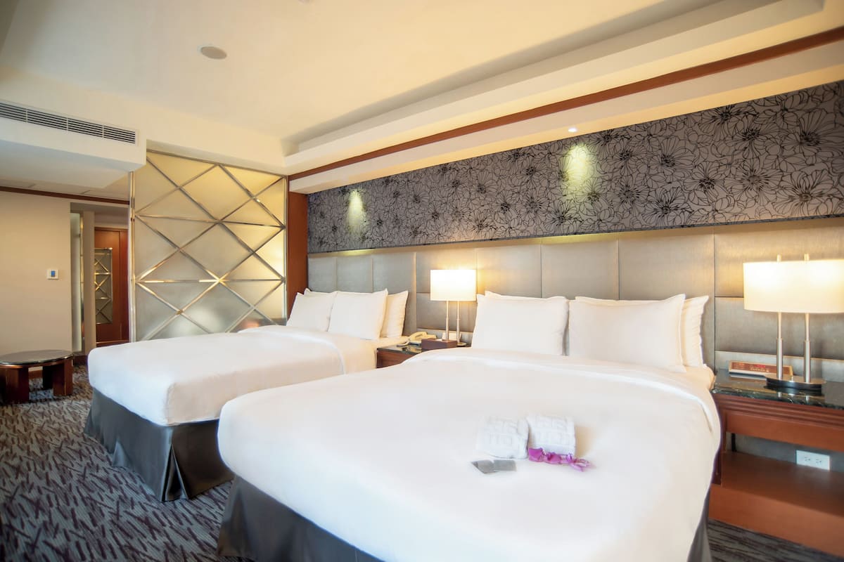 A hotel room with two beds, a large wall-mounted headboard, bedside lamps, and a carpeted floor.