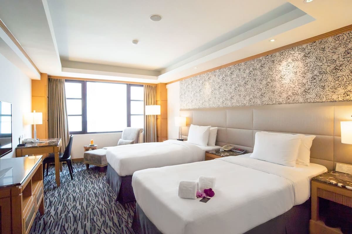 A hotel room with two beds, a desk, a chair, a TV, and a patterned wall.