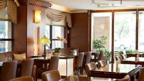 A hotel dining area with multiple tables and chairs, a potted plant, and a large window providing a view of the outdoors. - Hotel Dion (Taichung)