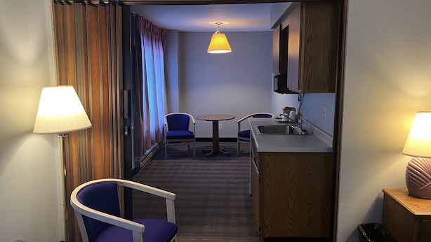A hotel room with a kitchenette, a dining area with two chairs, a small table, and a lamp. - The Ridge Hotel Portage (Portage)