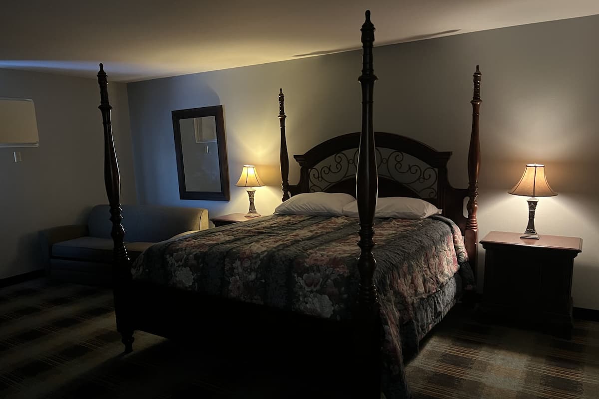 A four-poster bed with a floral bedspread, two bedside lamps, a chair, and a framed picture on the wall.