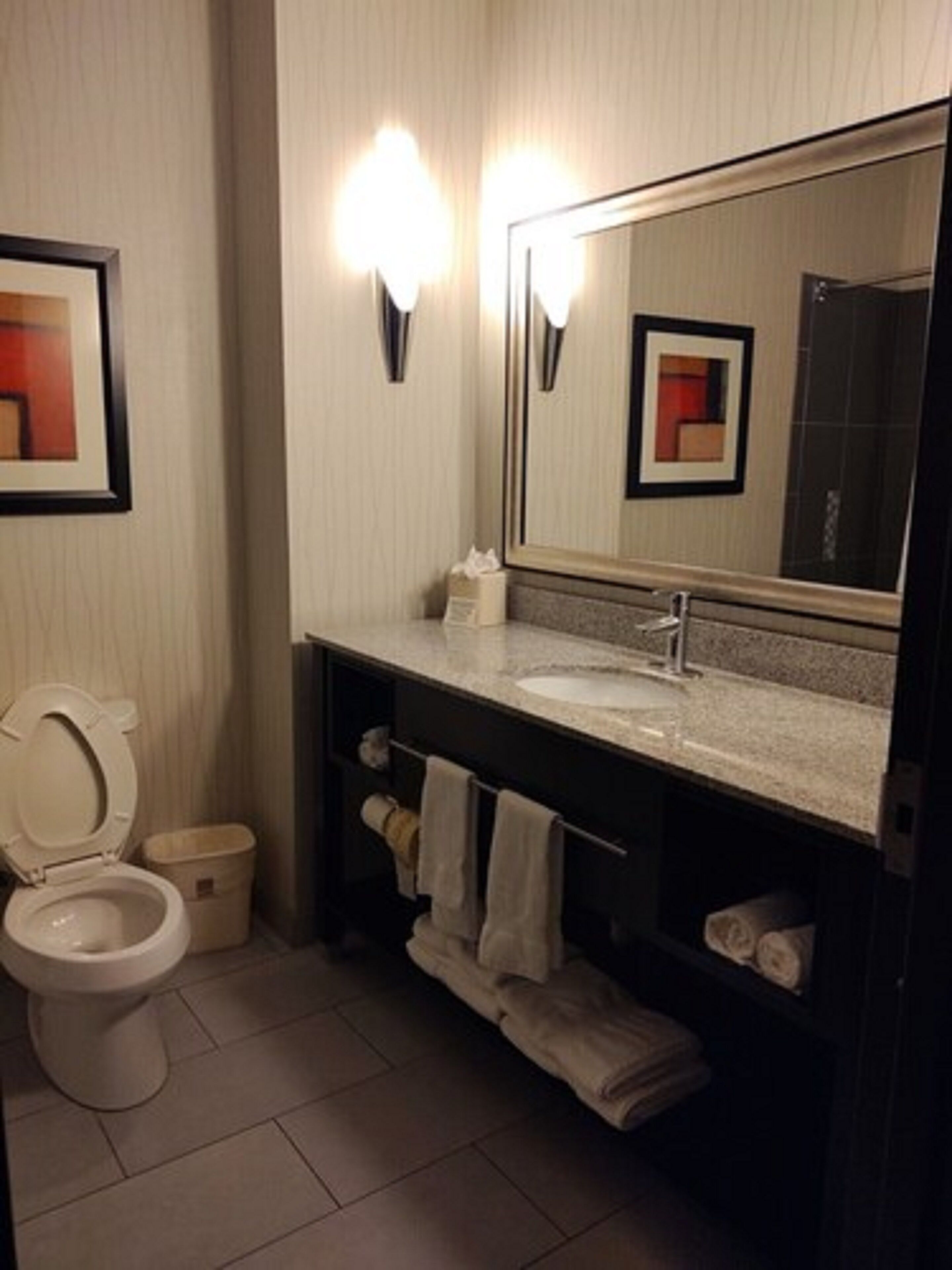 A modern bathroom with a toilet, sink, and mirror. There are towels and a trash bin.