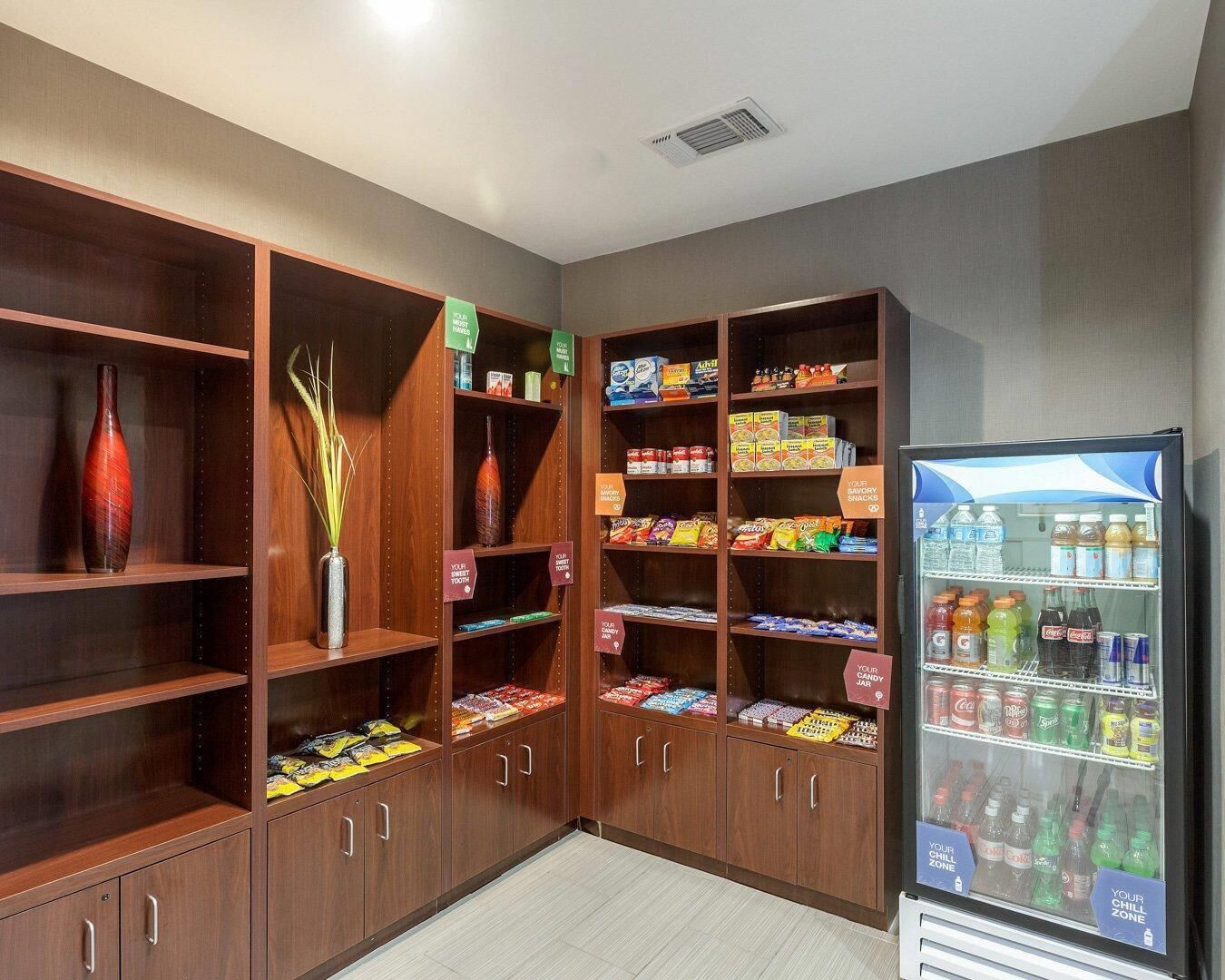 Habitación de hotel con estantes de madera llenos de aperitivos y un refrigerador que dice 'Tu Zona de Relax' con bebidas.