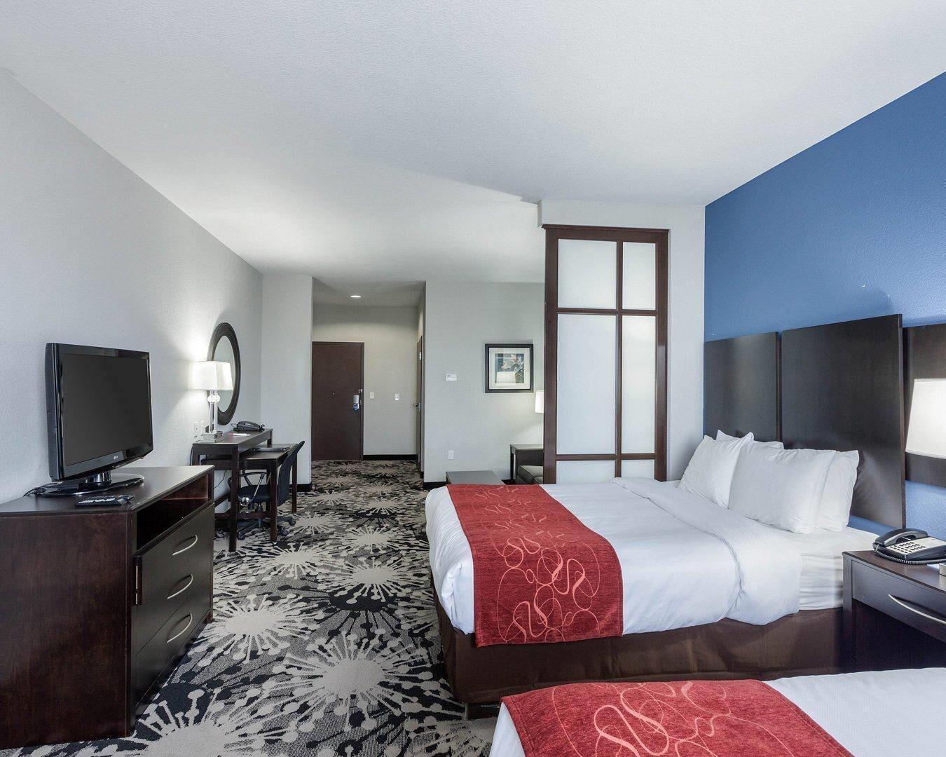 A hotel room with a bed, bedside table, TV, desk, chair, and a patterned carpet.