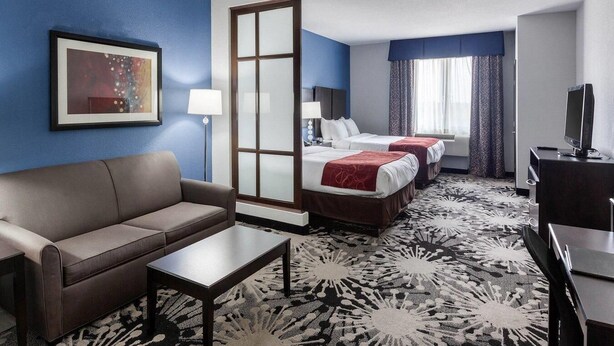 A hotel room with a sofa, two beds, a desk, a TV, and a large window with curtains.