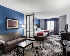 Ein Hotelzimmer mit Sofa, zwei Betten, Schreibtisch, Fernseher und einem großen Fenster mit Vorhängen.