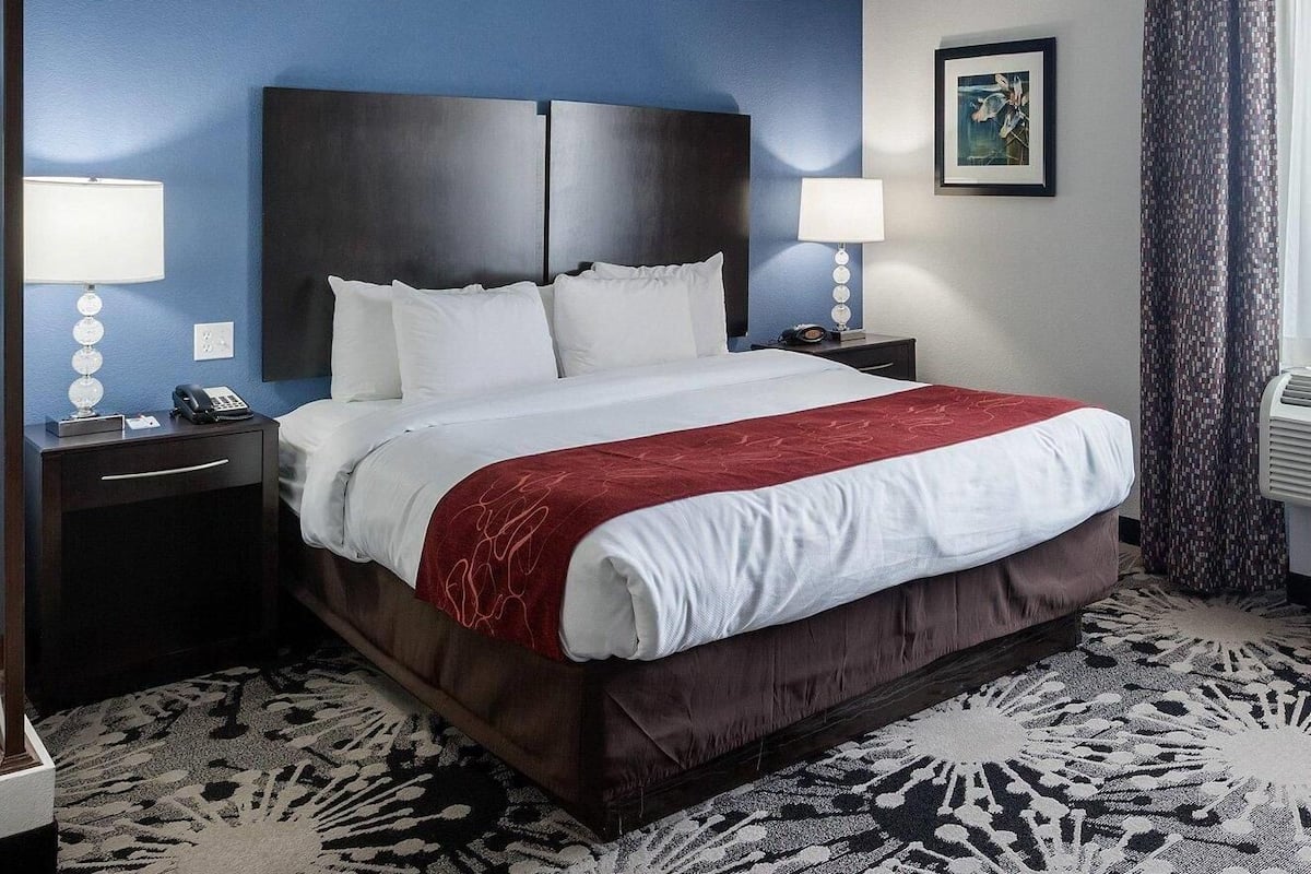 Une chambre d’hôtel avec un grand lit, deux tables de chevet, une petite table, une lampe fixée au mur, un tableau encadré et un tapis à motifs.