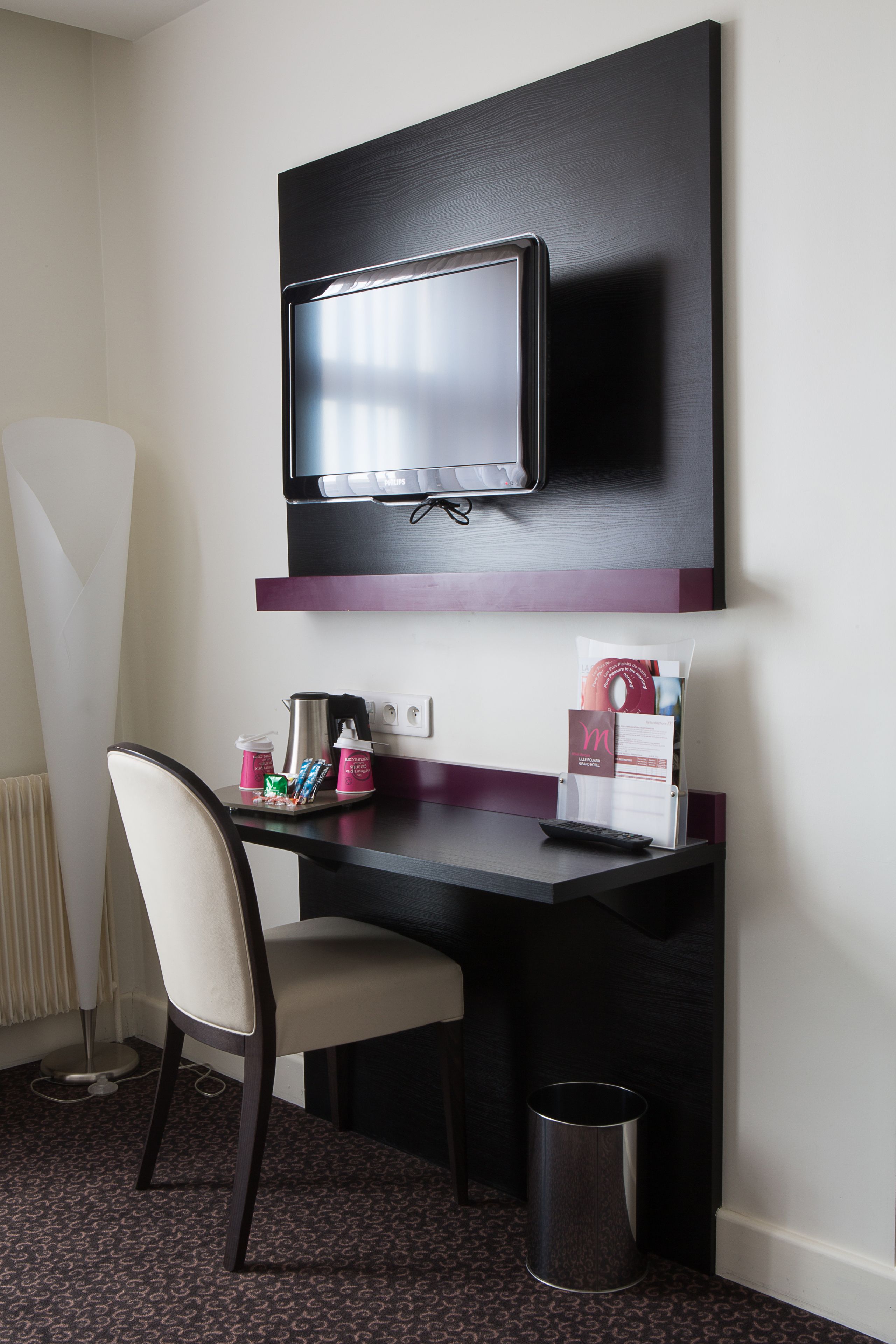 A hotel room with a desk, chair, TV, and a trash can.