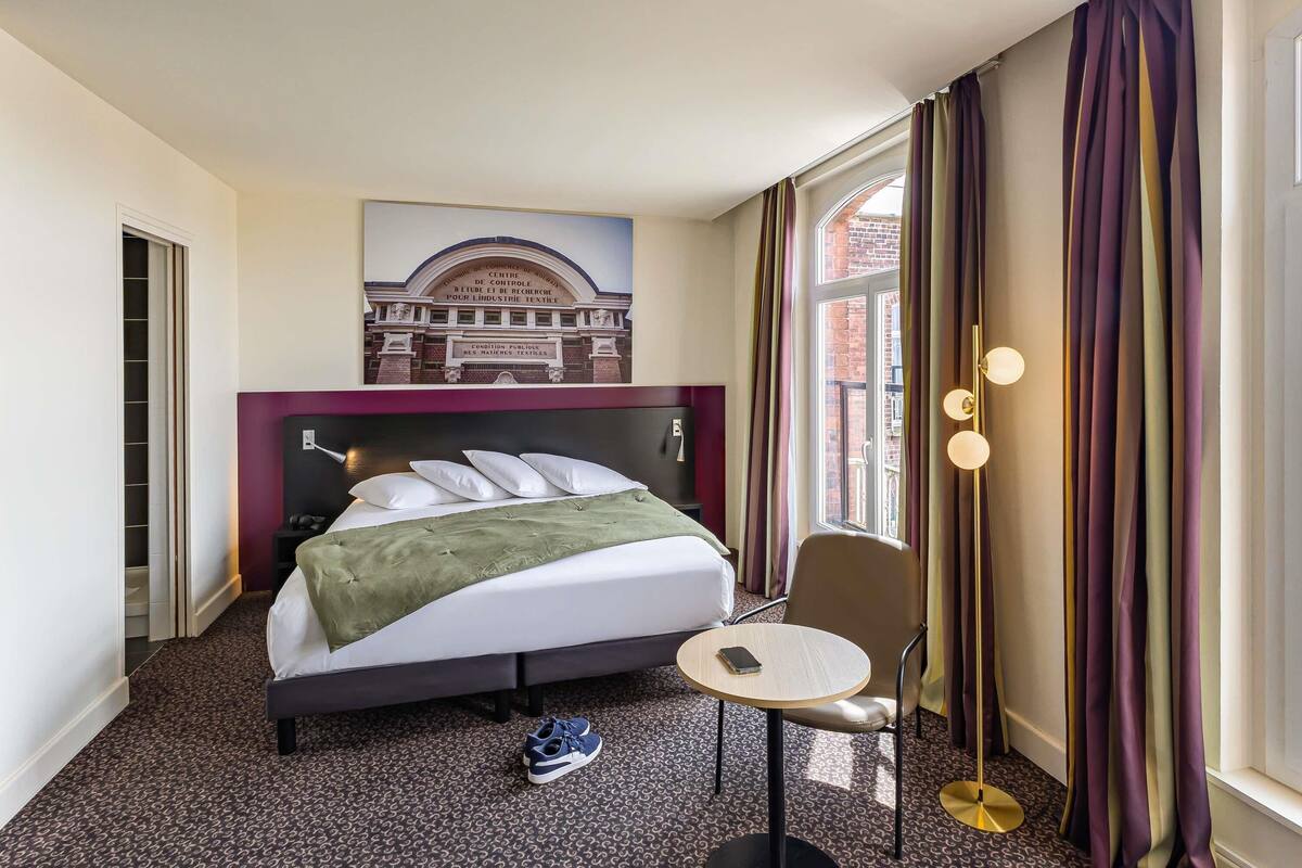 A hotel room with a bed, a small round table, a chair, a pair of shoes, and a window with curtains.