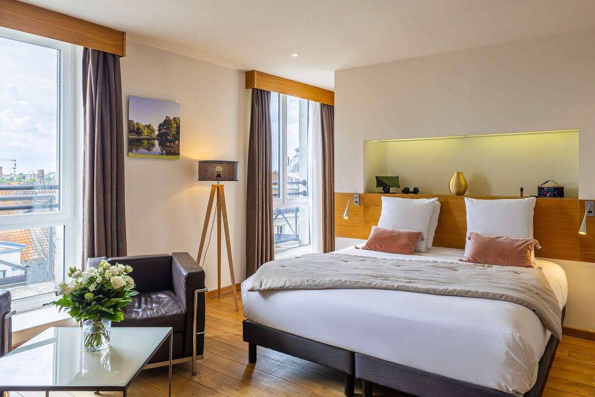 A hotel room with a bed, bedside table, chair, and a view of the city.