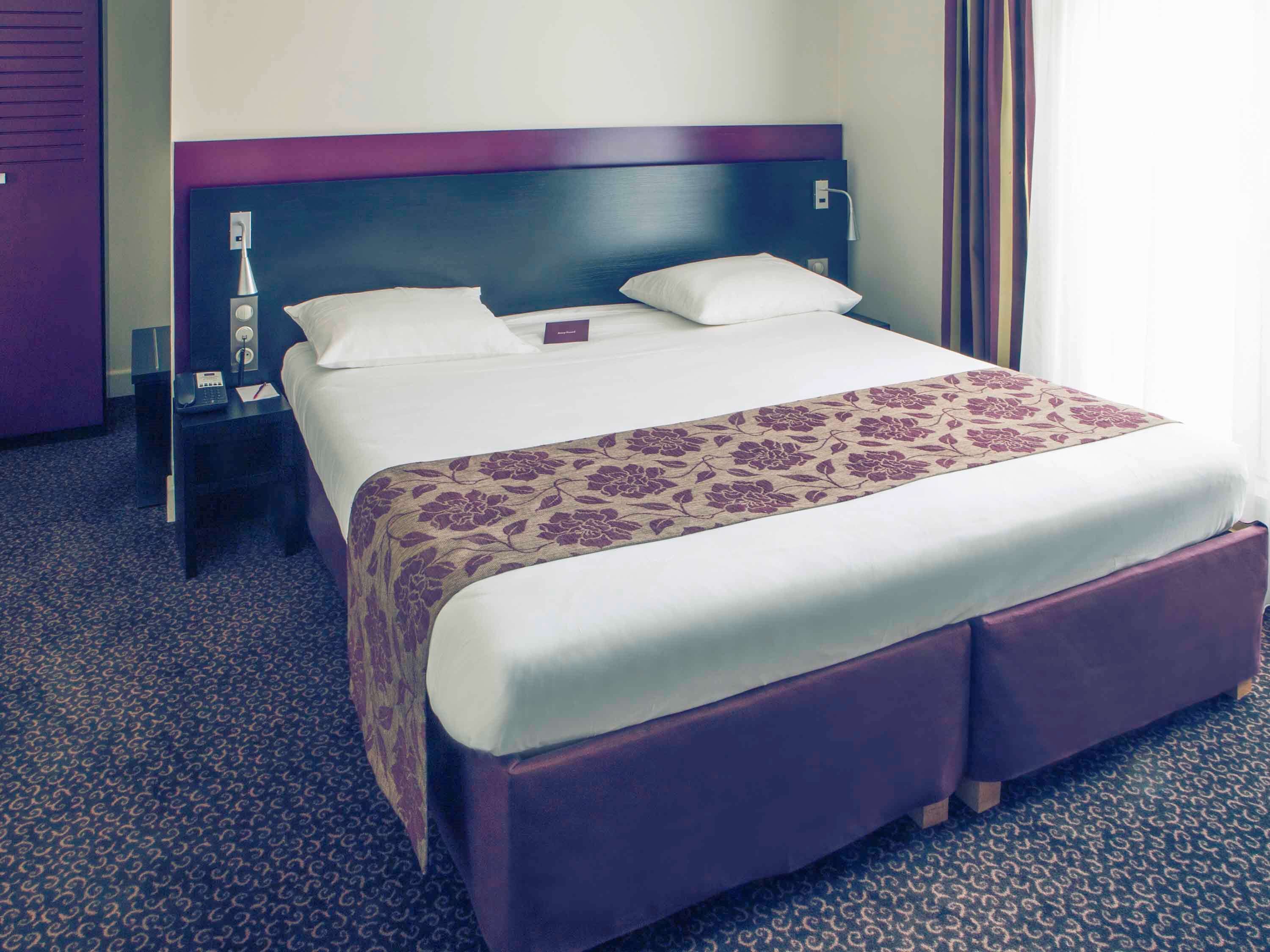 A hotel room with a large bed, a headboard, a bedside table with a lamp, and a carpeted floor.