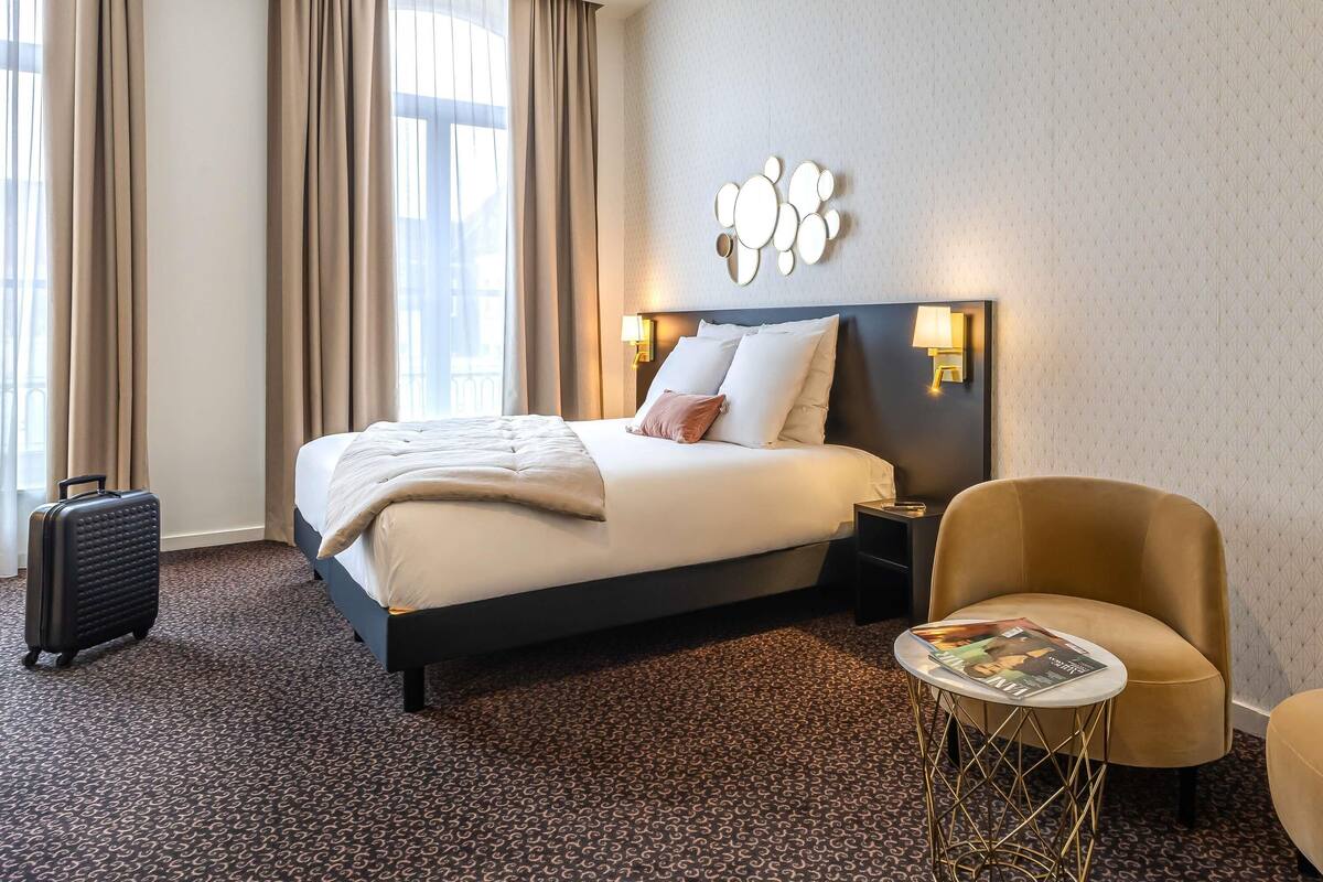 A hotel room with a bed, a chair, a small table, and a suitcase.