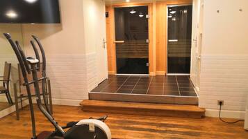 A home gym with a treadmill, stationary bike, and exercise equipment. There is a TV mounted on the wall and a wooden staircase leading to a dark room.
