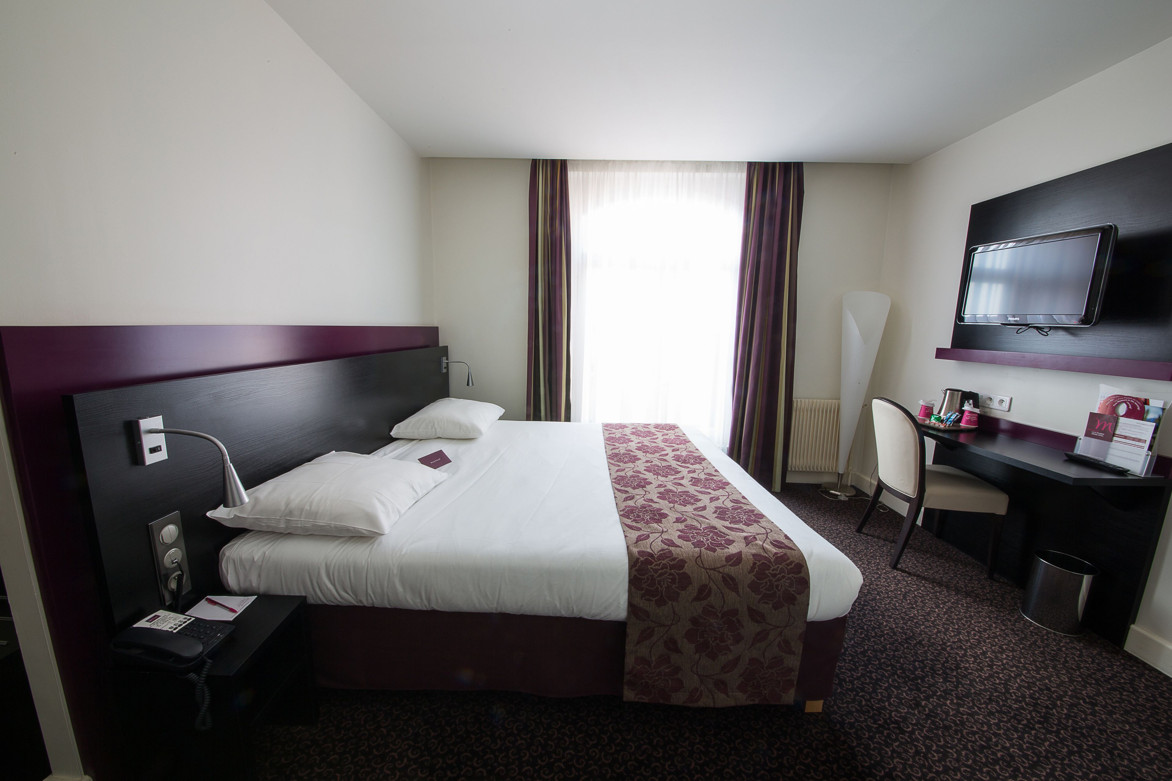 A hotel room with a bed, desk, chair, TV, and a window with curtains.