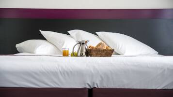A neatly made bed with white pillows, a basket of bread, a glass of orange juice, and a coffee pot.
